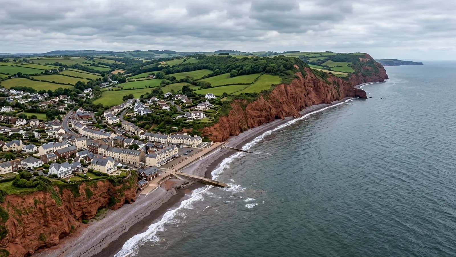 Red sandstone cliffs and a shingle beach on the East Devon coast with a small resort town of rendered and slate roofed buildings behind the sea front